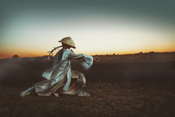 Blonde Asian woman with a long braid doing martial arts in a field in front of a sunrise while holding a wooden egg by Artmood Visualz